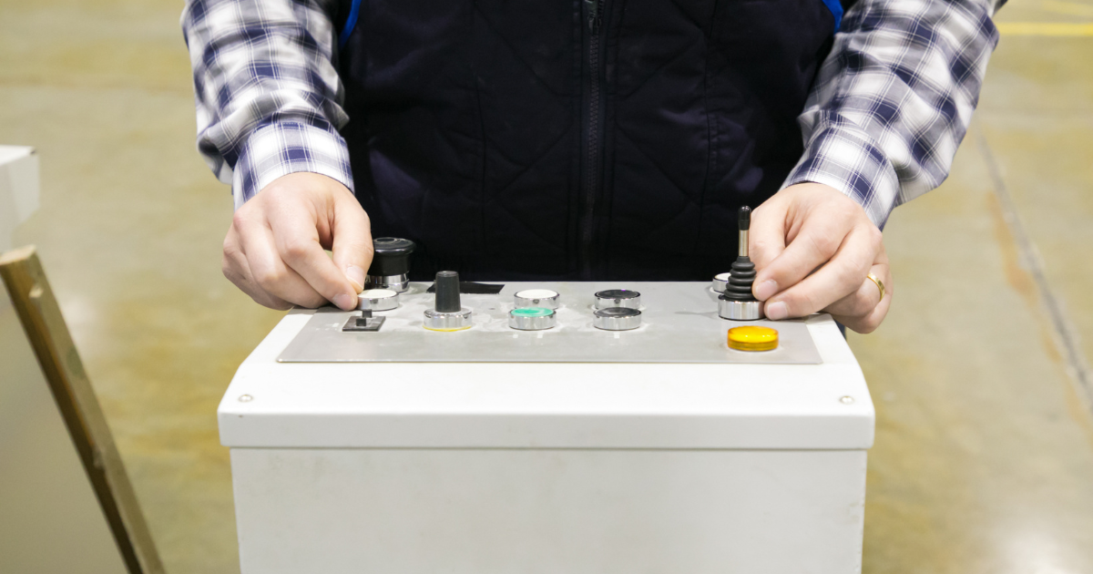 A person operating a control panel with various buttons and levers, wearing a plaid shirt and a dark vest.