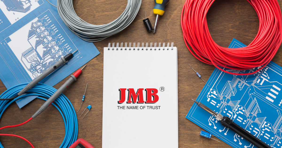 Electronics workshop with various components like wires, resistors, capacitors, and soldering tools. The JMB logo emphasizes their position as a trusted Fan Regulator Manufacturer in India.