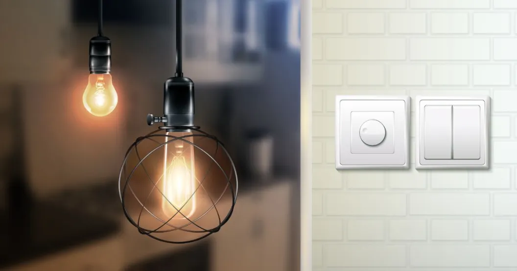 A close-up of a dimmer switch and light switches on a tiled wall, with vintage-style hanging light bulbs in the background. Featured by leading fan regulator manufacturers in India.